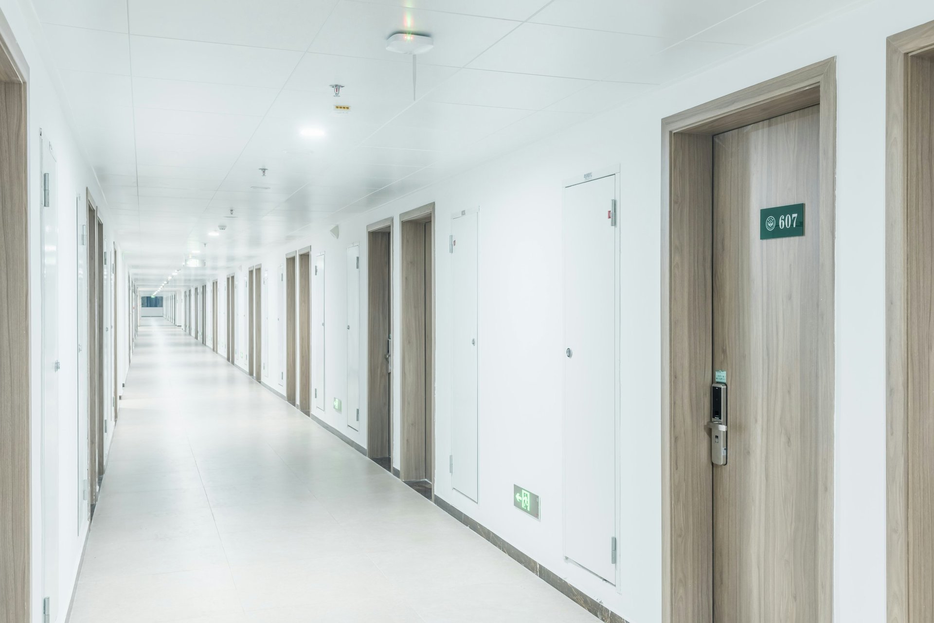 Long, modern hallway with white walls, multiple doors on both sides, and an exit sign visible at the end