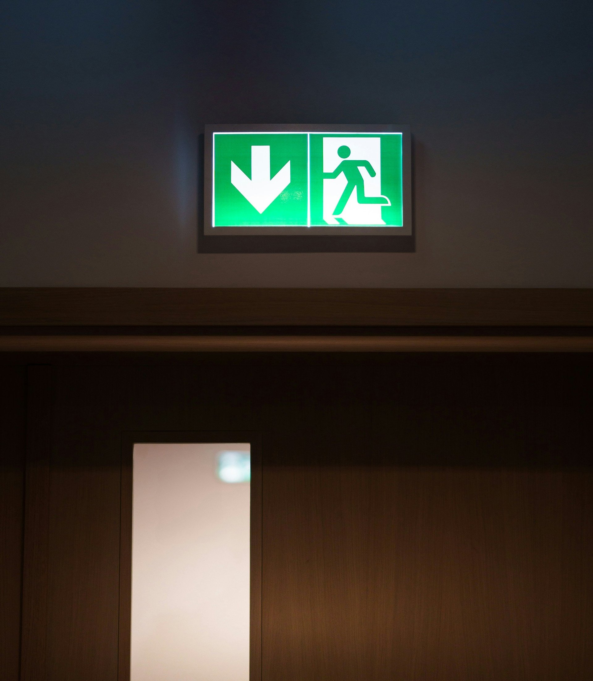 Green illuminated emergency exit sign showing downward arrow and running figure with doorway below